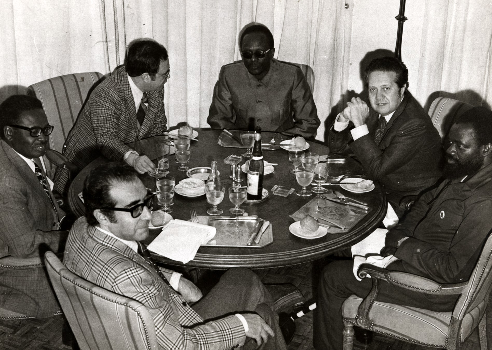 Almoço durante a cimeira entre Portugal e os três movimentos de libertação de Angola, no Alvor. Da esquerda para a direita, o ministro da Coordenação Interritorial Almeida Santos, o presidente do MPLA Agostinho Neto, o ministro Sem Pasta Melo Antunes, o presidente da FNLA Holden Roberto, o ministro dos Negócios Estrangeiros Mário Soares e o presidente da UNITA Jonas Savimbi, 12 de janeiro de 1975. Fotografia de C. Mota. Fonte: ANTT, SNI